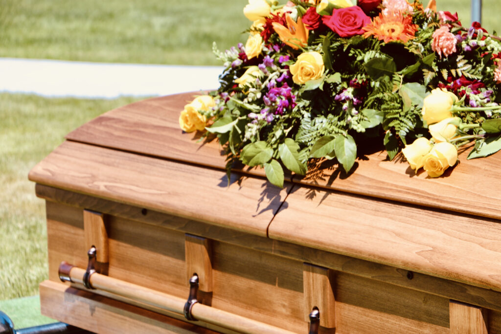 Wooden funeral casket with a vibrant floral arrangement on top, set outdoors on a grassy area, symbolising respect and remembrance.