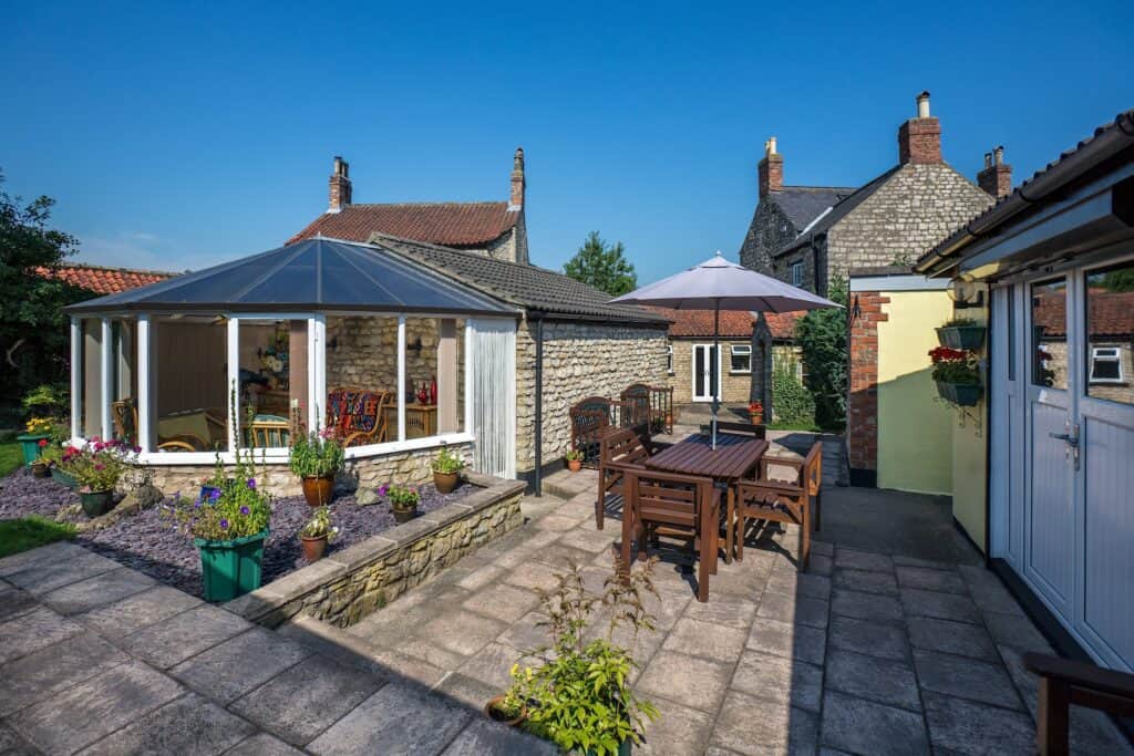 Bright outdoor garden patio with wooden furniture, potted plants, and a glass conservatory, showcasing a peaceful garden space for relaxation and outdoor dining.