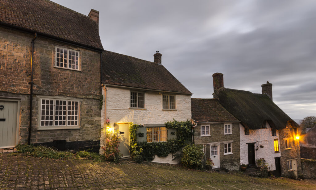 Charming historic cottages with traditional thatched roofs and stone walls, illuminated by warm evening lights, showcasing classic UK architecture.