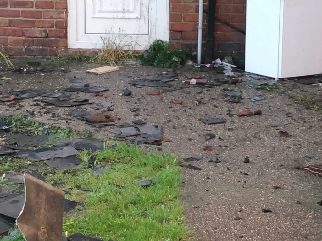 Broken slate roof tiles after storm damage outside property with brick wall and garden, urgent property repair and restoration needed.