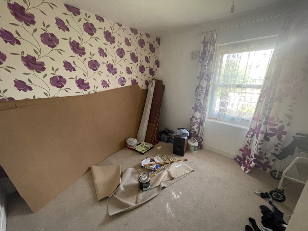 Cardboard panel leaning against the wall in a cluttered room with floral curtains and wallpaper, suggesting a property refurbishment or renovation.