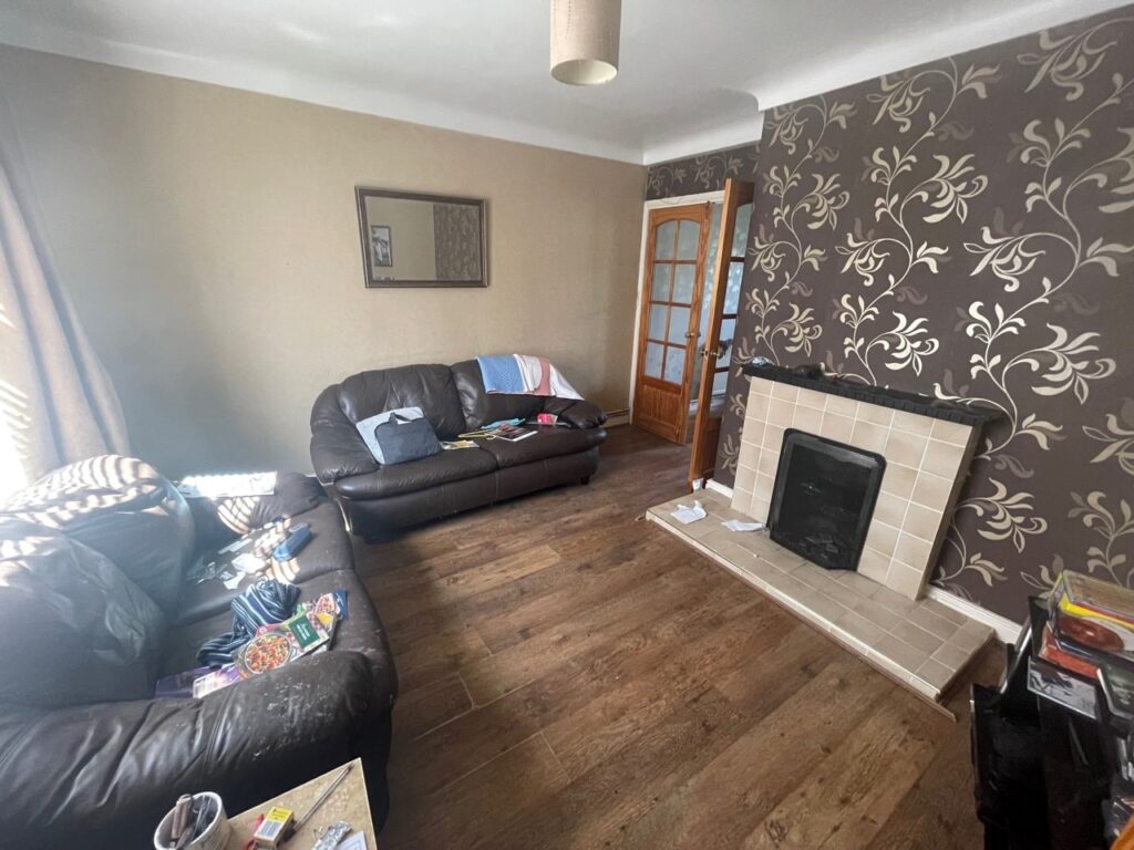 Cluttered living room with leather sofas, patterned wallpaper, and a fireplace, showcasing a typical home interior with scattered magazines and utensils for real estate or home improvement context.