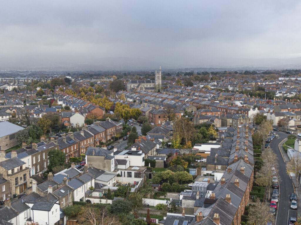 Aerial view of a dense urban neighbourhood in autumn, featuring rows of terraced houses and a distant church under an overcast sky.