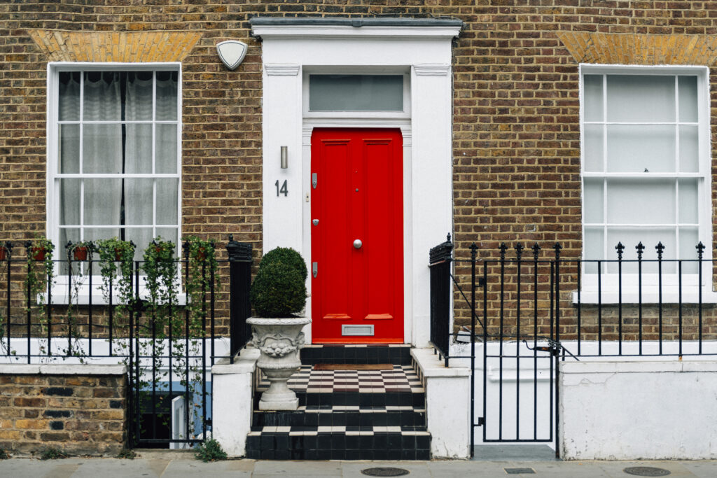 Red front door of a London townhouse, brick facade, two large white windows, black wrought iron fence, potted plants on porch.