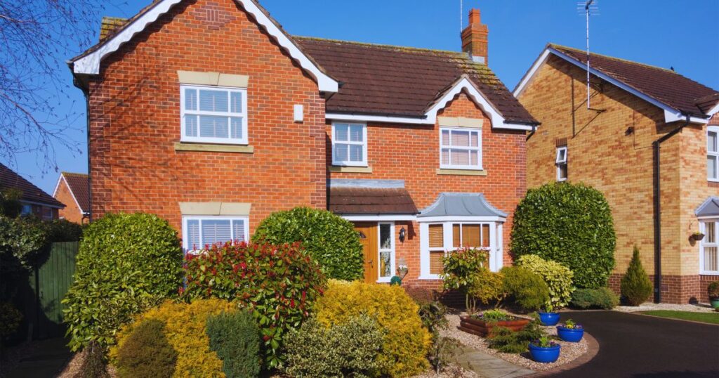 Charming red-brick home with pitched roof, white window frames, lush garden with vibrant shrubs, under a clear blue sky.