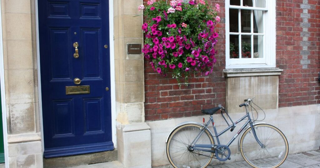 Bright blue front door with brass hardware, vibrant pink flowers in hanging basket, and vintage bicycle outside a brick house, showcasing property enhancement and curb appeal.