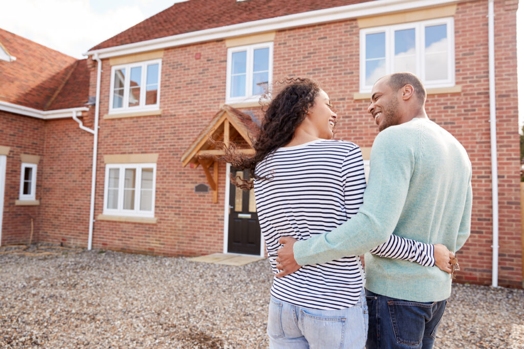 Happy couple embracing outside new red-brick house, man holding keys, symbolising new beginnings and home ownership.