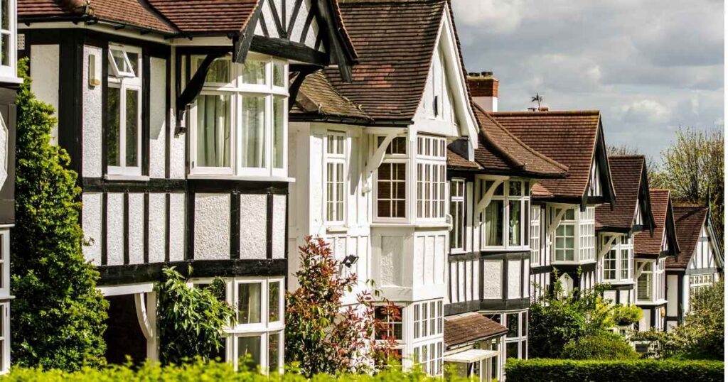 Victorian-style terraced houses with black timber framing and white stucco exterior, featuring bay windows and sloped roofs in a lush green neighbourhood.