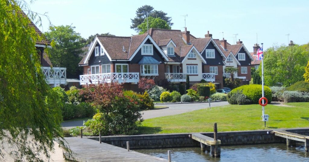 Luxurious residential property with waterfront view and landscaped gardens, featuring a traditional British style with red brick facade, gabled roofs, and white balcony railings.