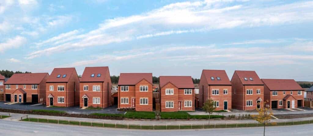 Modern new-build houses with red brick exteriors and pitched roofs, some with "Sold" signs, in a residential development near a main road, showcasing property sales and estate agency services.