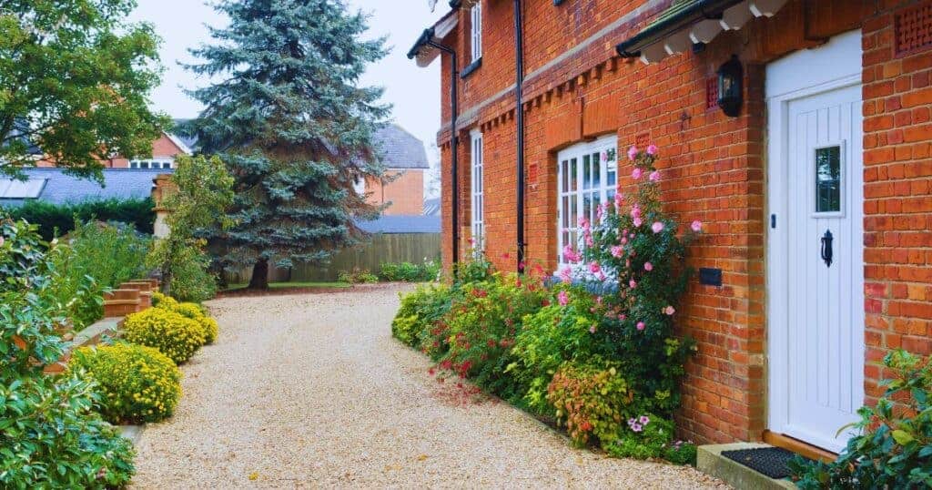 Bright red brick house with white front door and vibrant pink garden flowers, surrounded by lush greenery, gravel pathway, and mature trees in a peaceful residential setting.