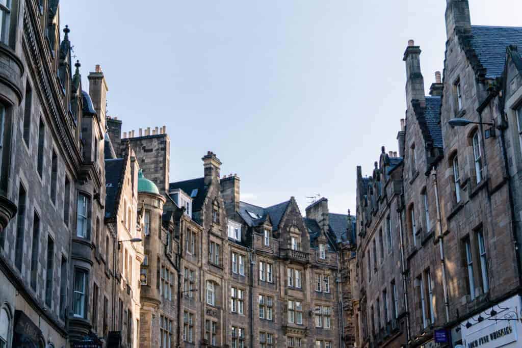 Historic stone buildings with gabled roofs line a narrow street under a clear blue sky, suggesting a traditional European city.