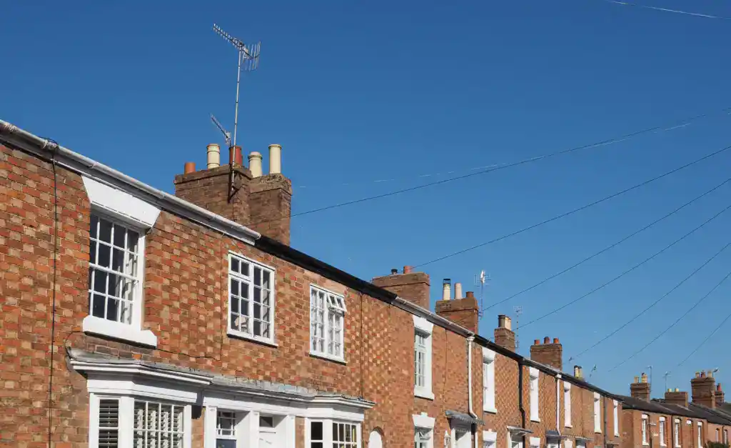 Row of traditional British terraced houses with brick walls, white windows, and chimneys against a clear blue sky.