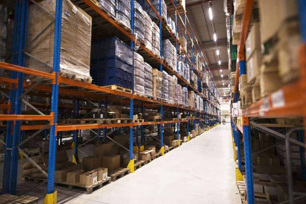 Warehouse aisle with tall shelves stacked with pallets of boxed goods, lit by bright overhead lights, creating an organised space.