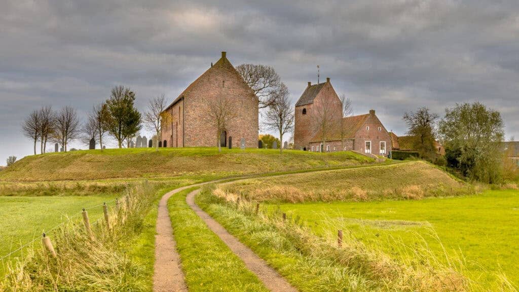 Ancient brick church on a green hilltop with a winding dirt path leading up to it; surrounded by trees and lush grass, under a cloudy sky, showcasing historic architecture and scenic rural landscape.