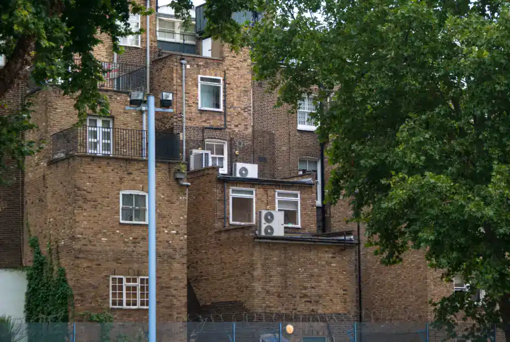 Historic brick apartment building with multiple air conditioning units, surrounded by lush green trees and a tall street lamp.