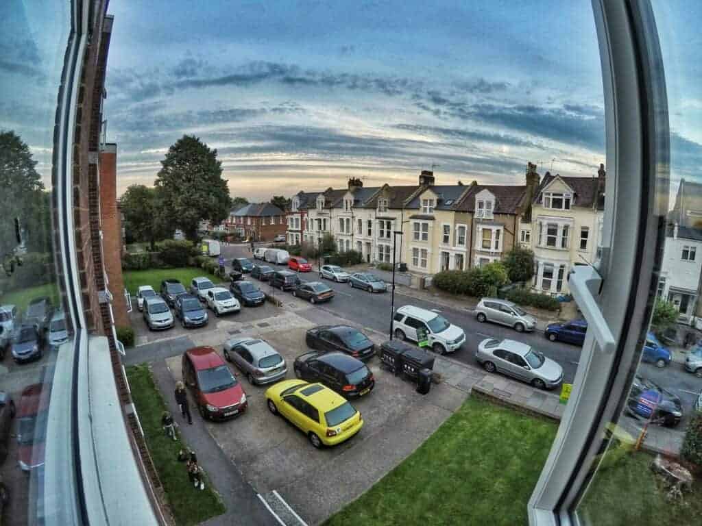 Street view from window showing parked cars, colourful houses, tree, and cloudy sky during sunset in residential area.