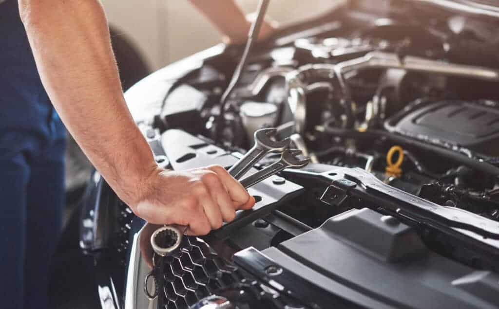 Mechanic's hand holding spanners near open car bonnet, likely performing engine maintenance or repair work.