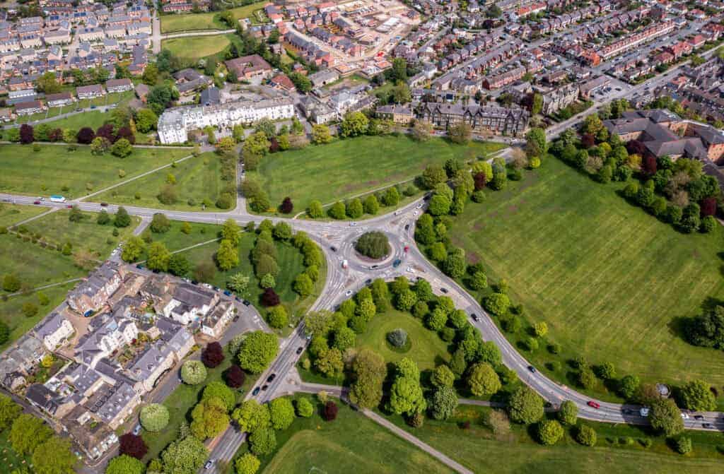 Aerial view of a roundabout in a suburban area surrounded by green parks and residential buildings, showcasing a well-planned landscape with trees and roads for an enhanced neighbourhood environment.