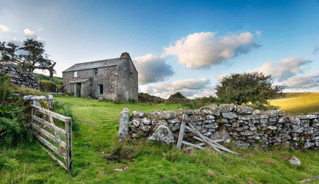 Rustic stone cottage in rural countryside with old stone wall and wooden gate, under a blue sky with clouds, showcasing traditional UK country property.