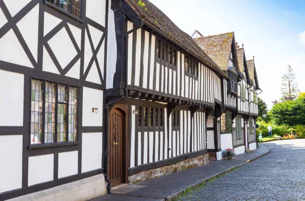 Charming Tudor-style house with black timber framing and white walls, situated on a cobbled street surrounded by lush greenery, showcasing traditional architecture in a serene environment.