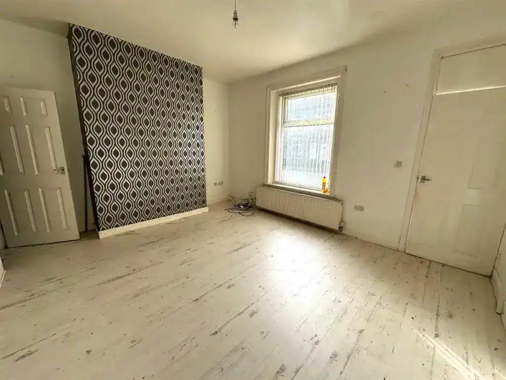 An empty room featuring a patterned accent wall, a large window allowing natural light, and white wooden flooring, ideal for a budget-friendly property in England.