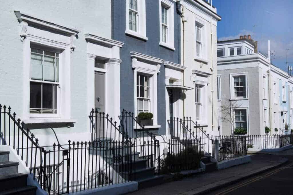 Bright pastel-coloured terraced houses in a seaside town, showcasing classic British architecture with white-framed windows, iron railings, and steps leading to front doors, under a clear blue sky.