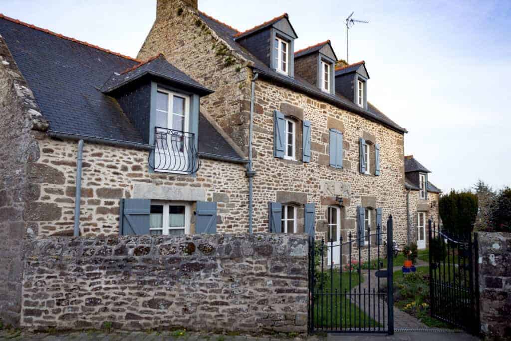 Charming traditional stone house with blue shutters, lush garden, and black wrought iron gate, ideal for property buyers seeking historic homes with modern upgrades and excellent street appeal.