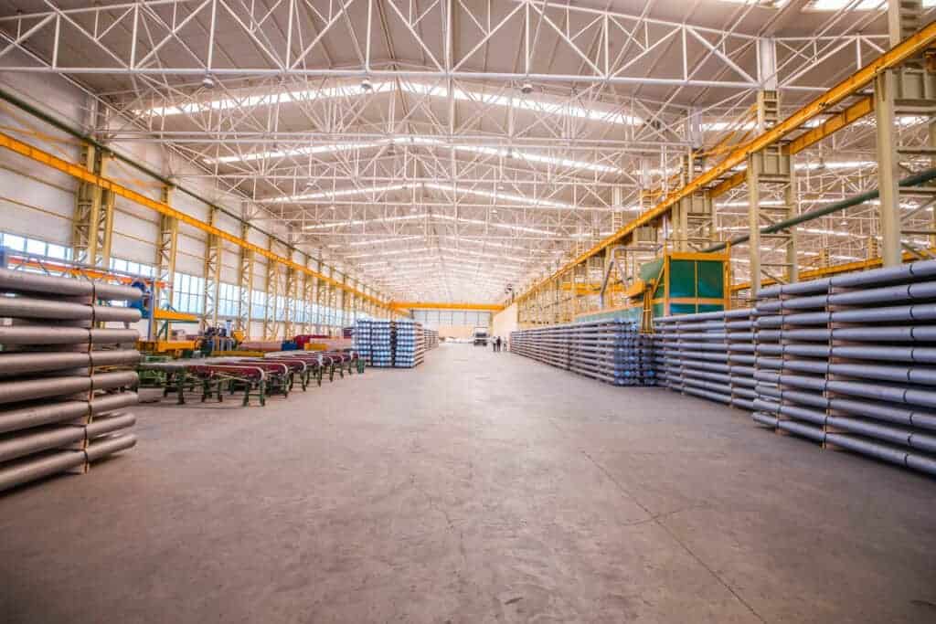 Spacious industrial warehouse with stacked metal pipes, high ceilings, and overhead cranes, showcasing a large storage area.
