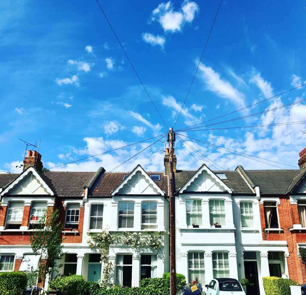 Row of traditional terraced houses with white facades and pitched roofs under a bright blue sky, featuring overhead power lines and a peaceful residential street scene.