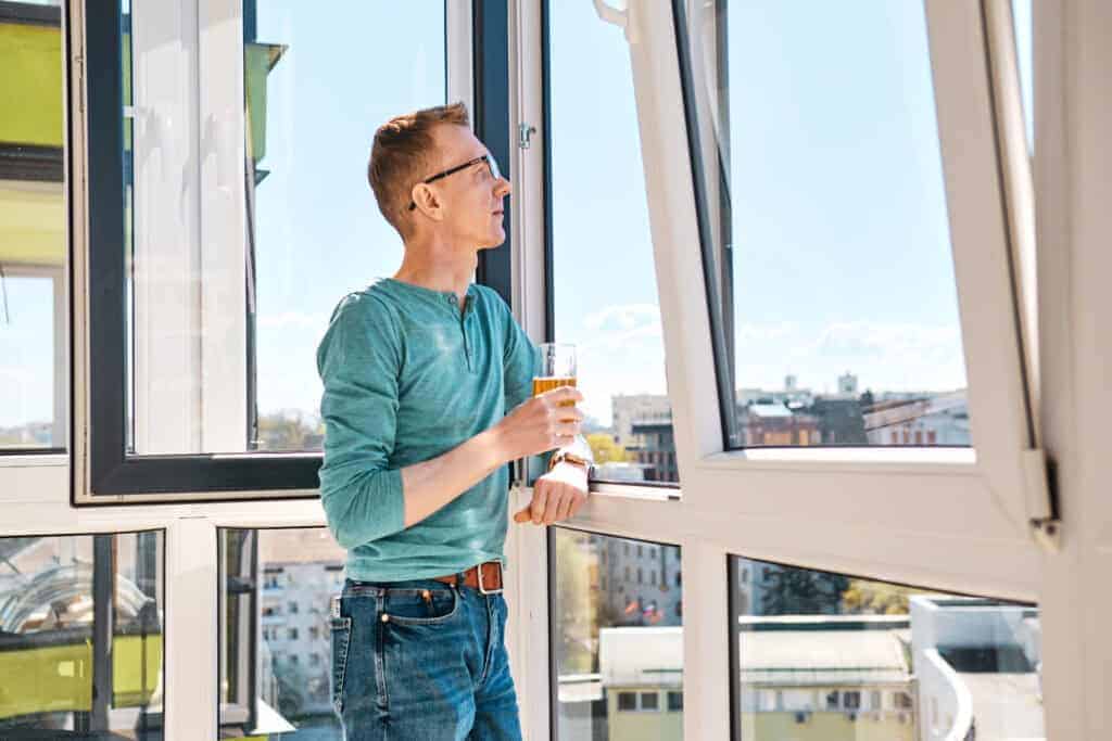 A man standing by an open window holding a glass of orange juice, overlooking a cityscape, representing home improvement and certification processes.