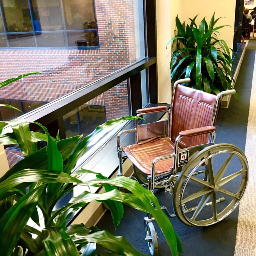 An image of a wheelchair positioned next to a large window with a view of a brick building, surrounded by green plants, highlighting accessibility and comfort for wheelchair users.