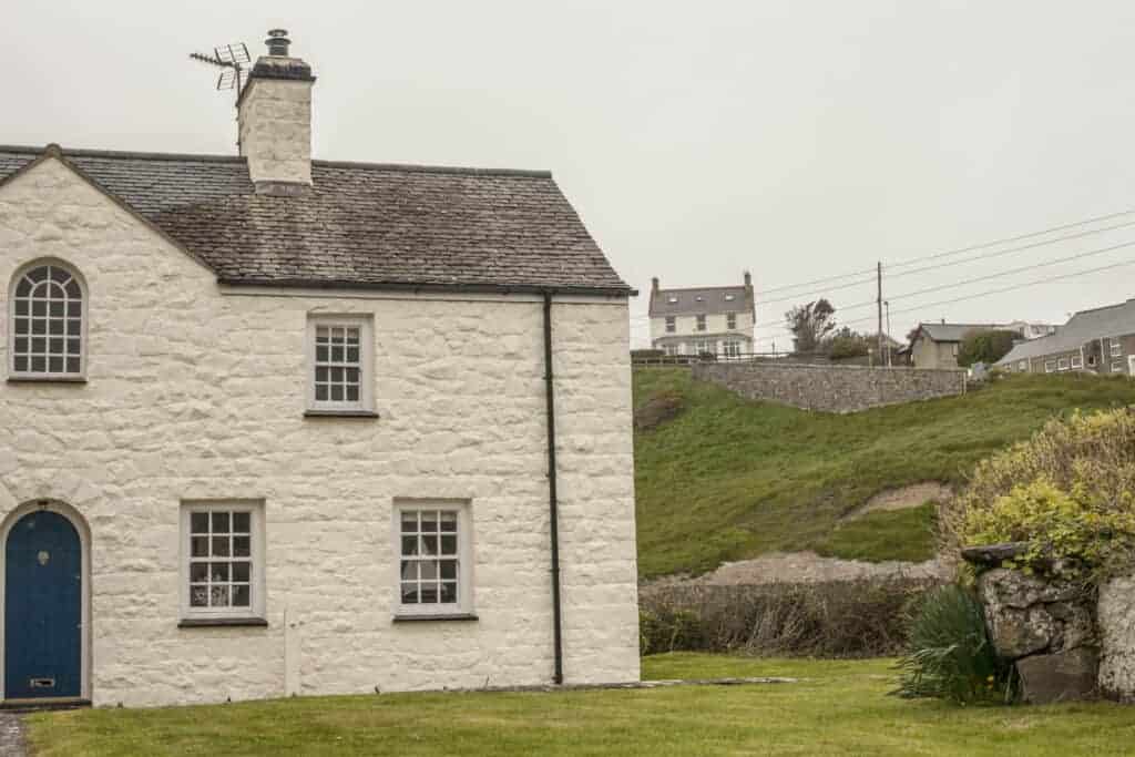 Charming white stone cottage with traditional windows and a blue door, set amidst lush green gardens and hillside background, capturing classic UK countryside architecture.