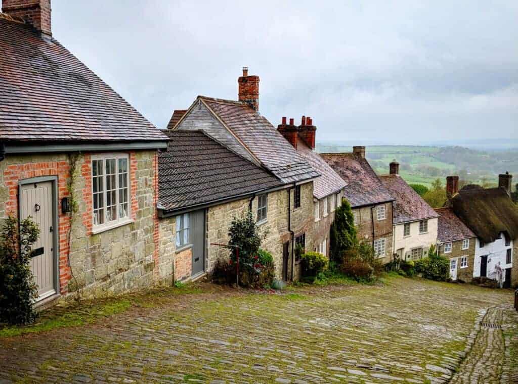 Traditional English village houses with stone and brick exteriors, cobbled street, overcast sky, scenic countryside backdrop, charming historic homes, rustic village scene, quintessential UK rural architecture.