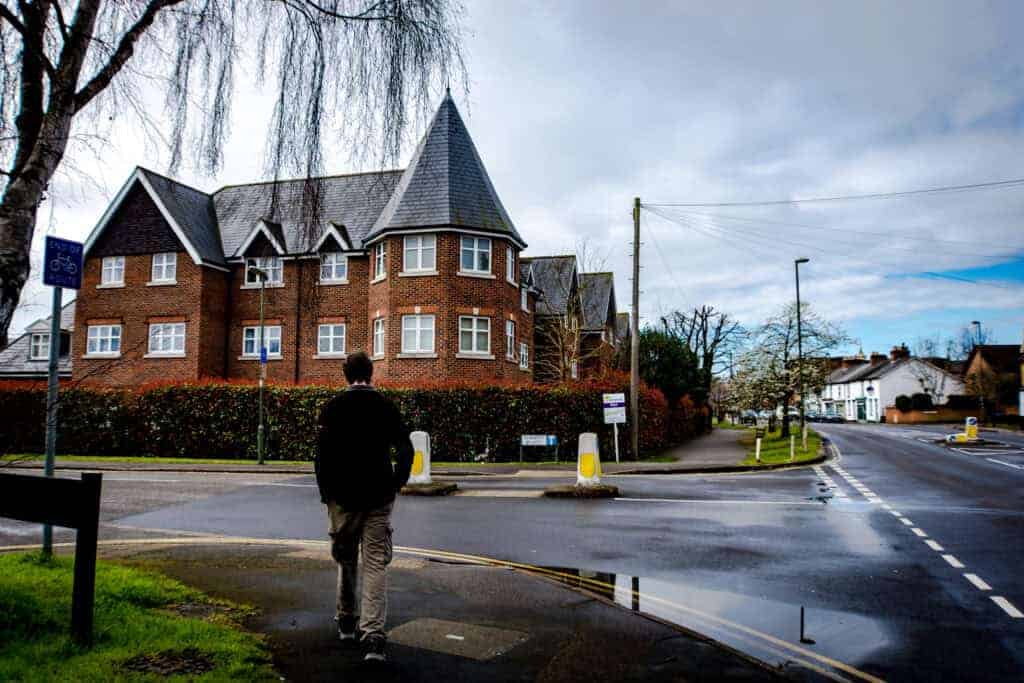 A person walking past a large, traditional brick house on a quiet street, illustrating online property selling.