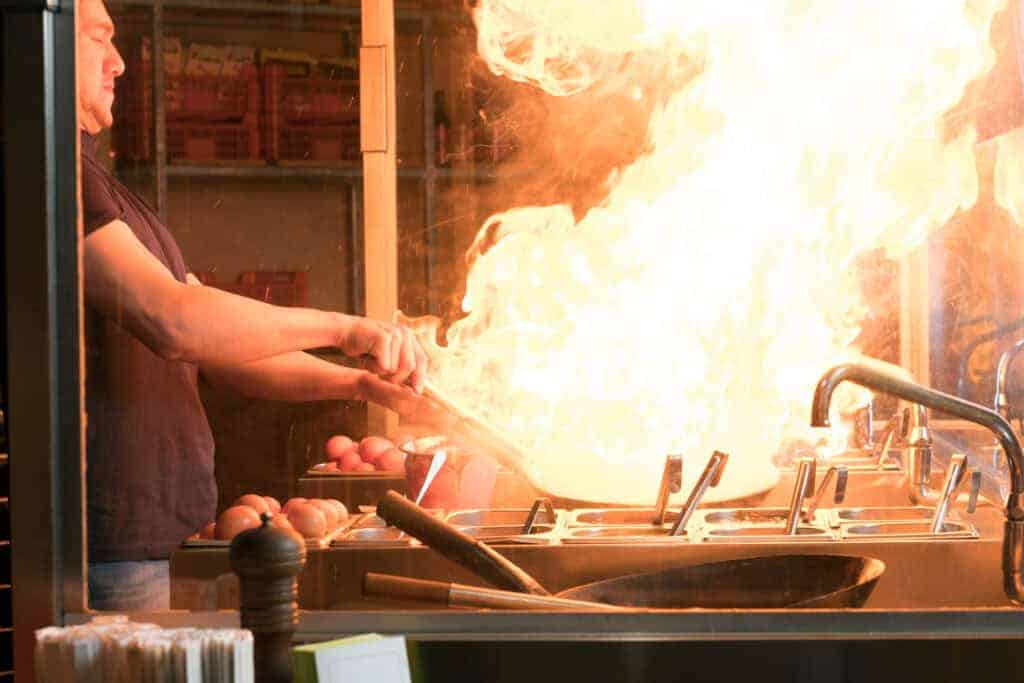 Chef flamb&eacute;ing in a kitchen, creating large flames over a stove, surrounded by pots and ingredients, with bright fire illumination.