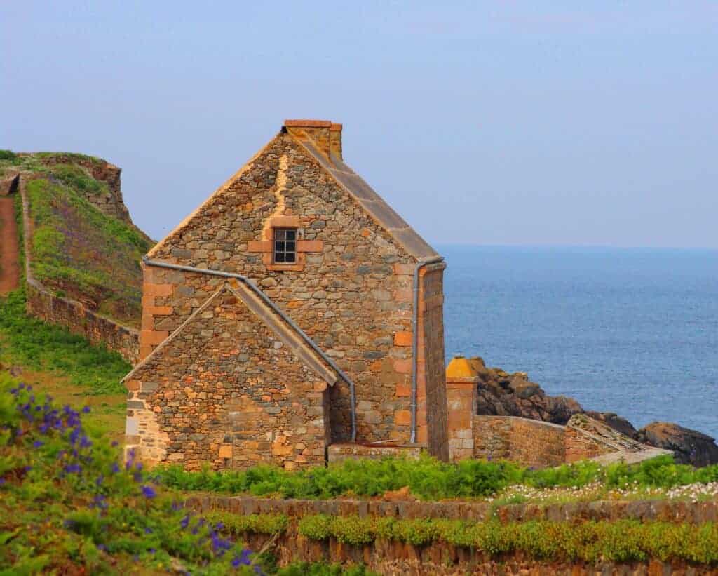 Charming stone cottage on cliffs overlooking the sea, nestled in lush greenery with colorful flowers, ideal for coastal property investment and seaside property management.