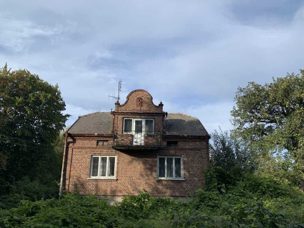 Old brick house with a small balcony, surrounded by greenery, under a blue sky with light clouds - Property Saviour specialist in property restoration and renovation.