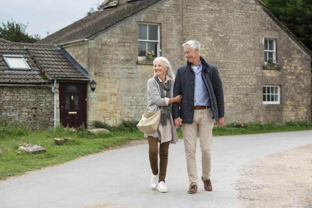 Older couple walking hand-in-hand near a rustic stone house, symbolising homeownership and retirement planning.