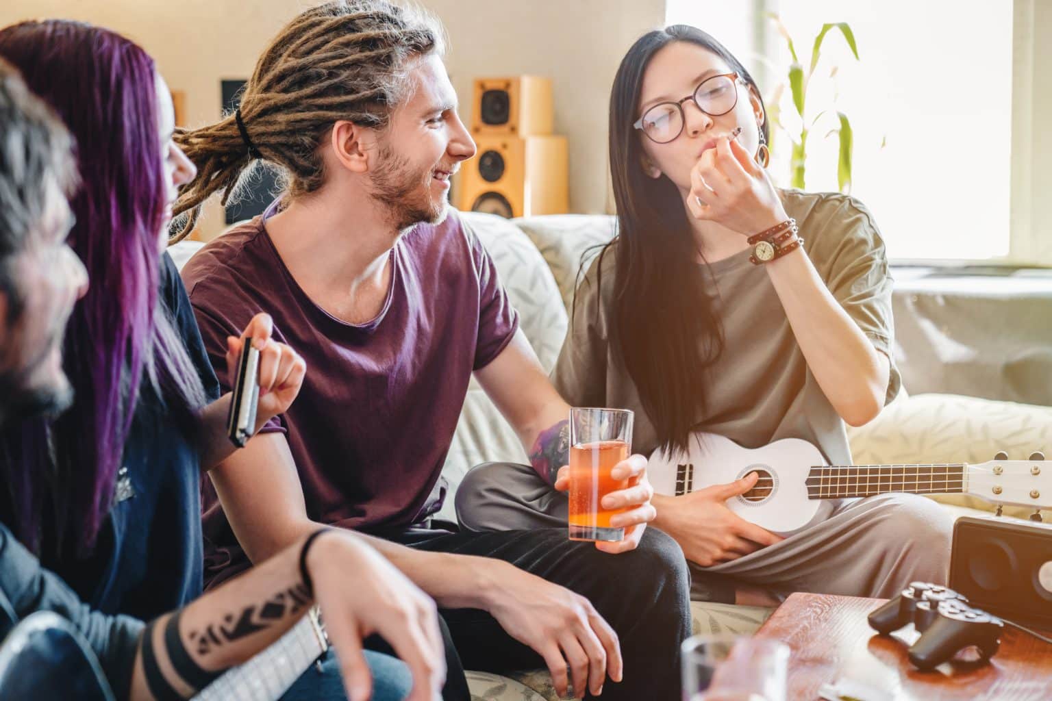 Group of friends relaxing, one with dreadlocks holding a drink, another playing a harmonica, and a woman with a ukulele chilling.