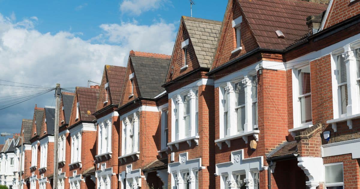 Row of traditional British terraced houses with red brick, white trim, gabled roofs, and chimneys under a partly cloudy sky.