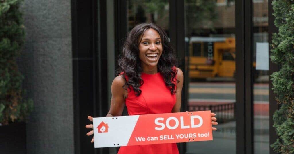 A woman holding a "SOLD" sign outside a property, smiling confidently, representing successful property sales and expert assistance in assisted sales.