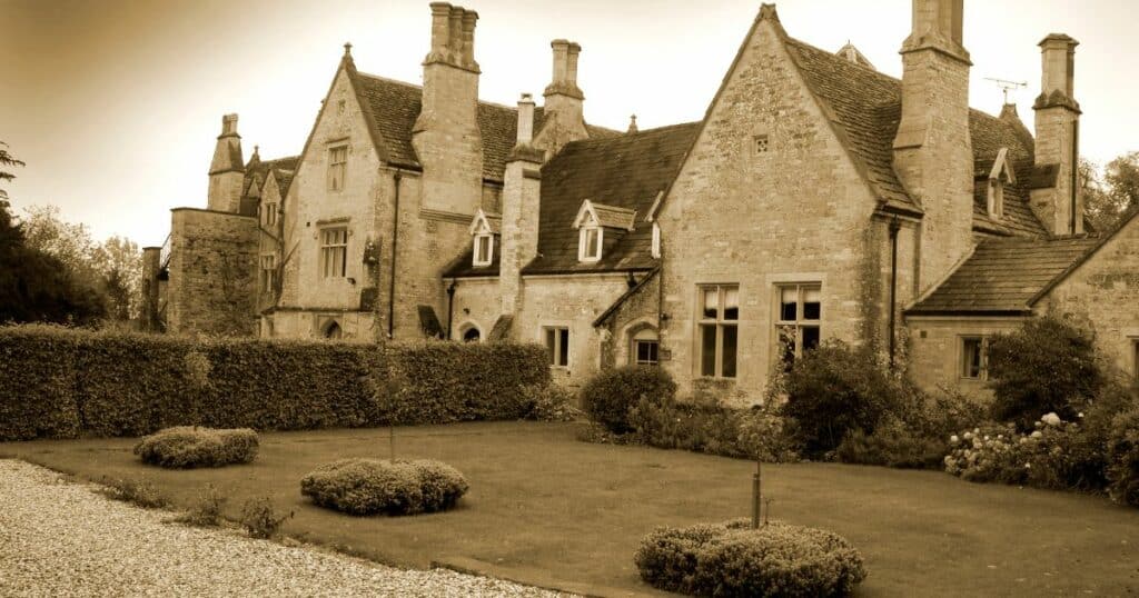 Sepia-toned photo of a large, historic stone manor house with gabled roofs, tall chimneys, and a well-kept garden in front.